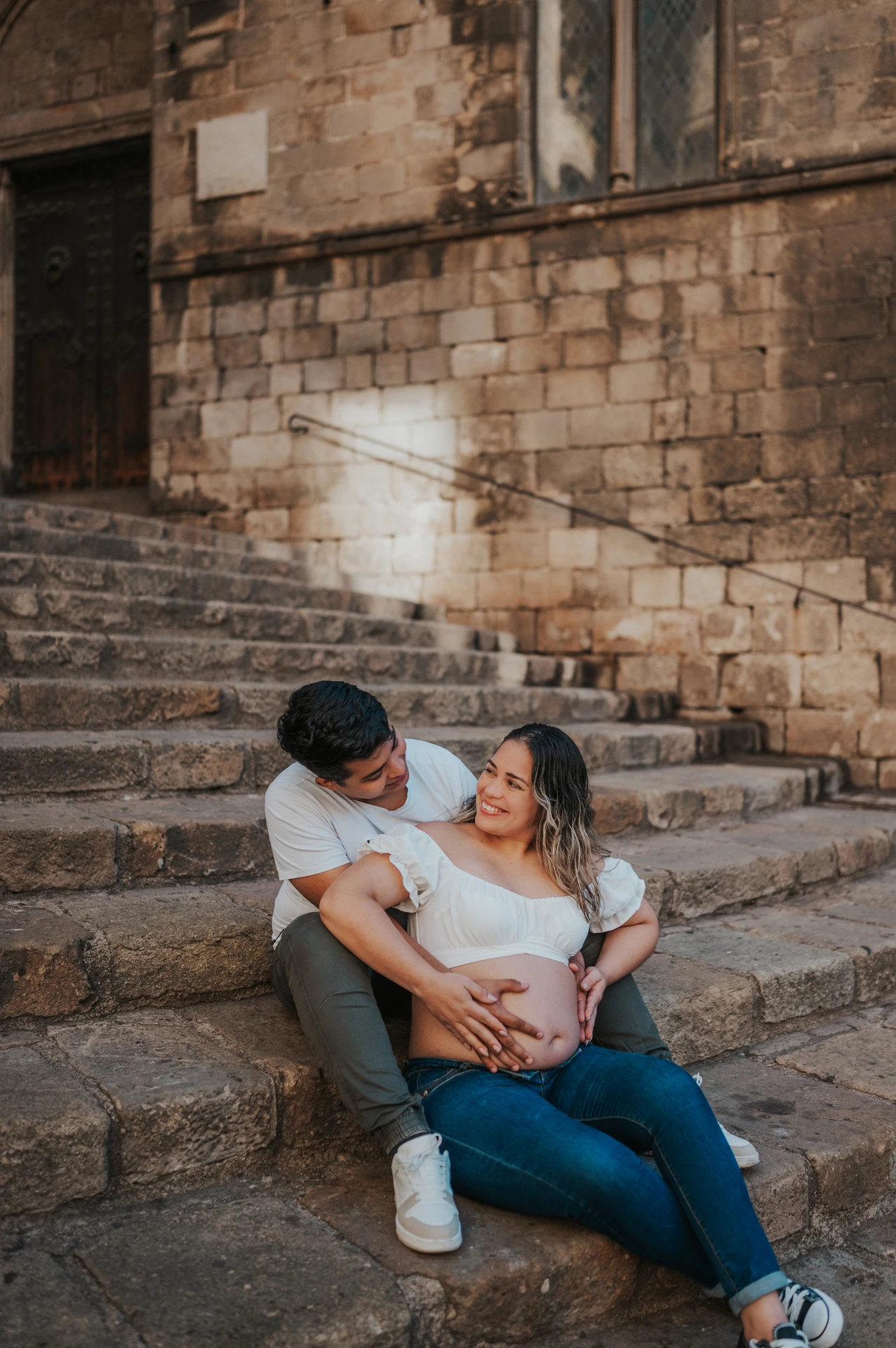 The couple sitting together on the stone steps with their hands resting on her bump during their maternity photoshoot in Barcelona.
