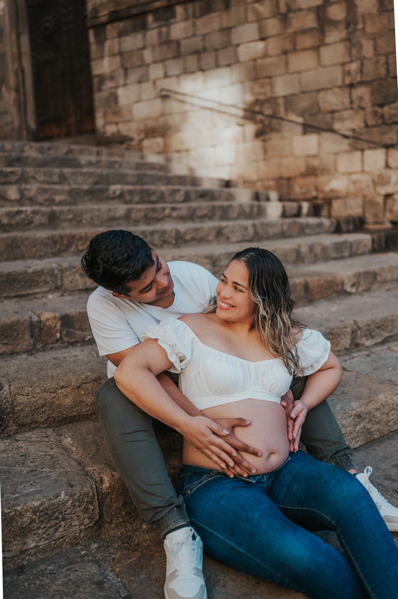 The couple sitting together on the stone steps with their hands resting on her bump during their maternity photoshoot in Barcelona.