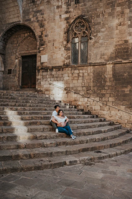 A wide portrait on the stone steps | Barcelona maternity photoshoot A wide view of the couple sitting on the steps beneath old stone walls and carved windows during their maternity photoshoot in Barcelona.