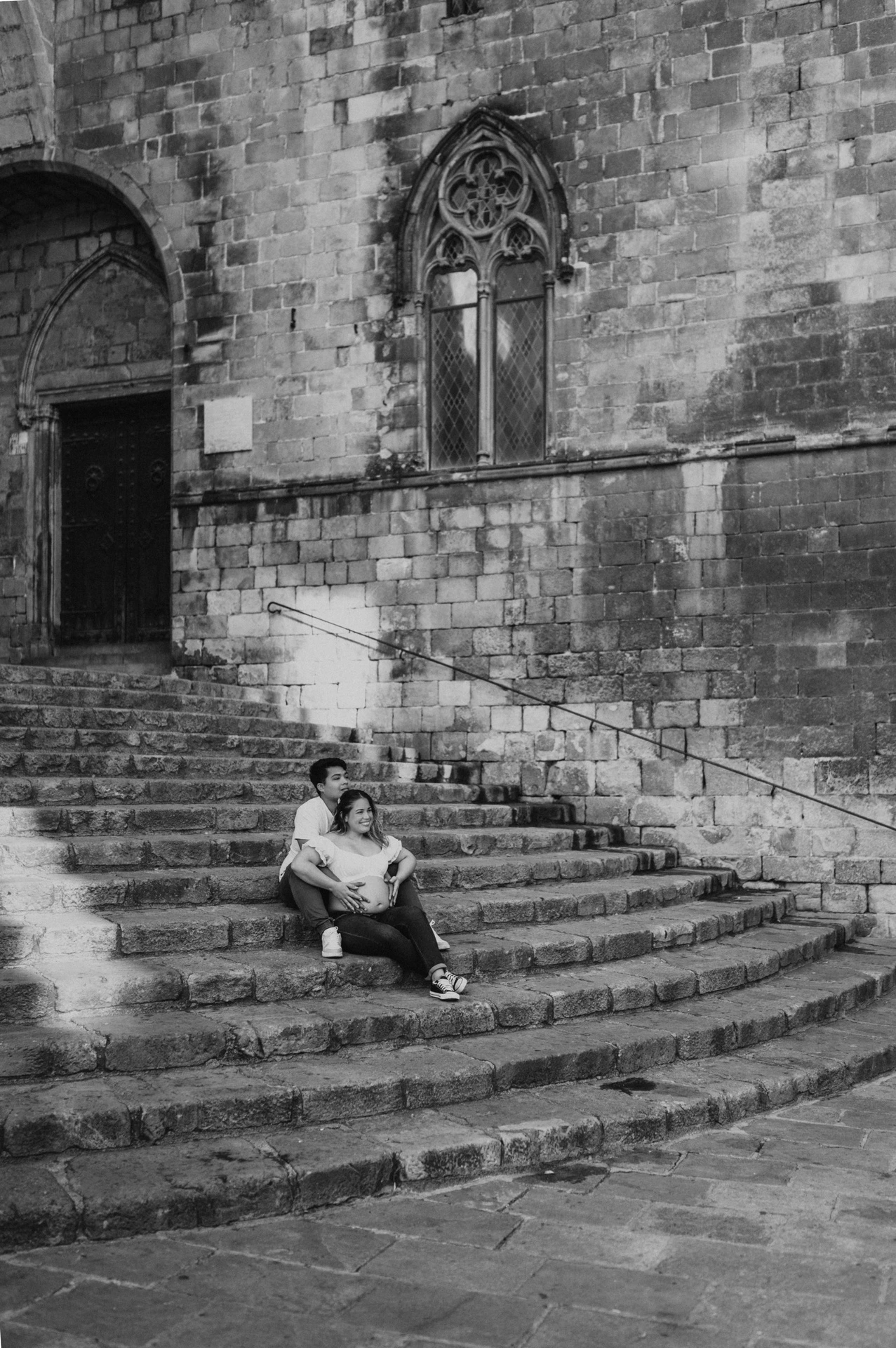 Black-and-white photograph: a wide view of the couple sitting on the steps beneath old stone walls and carved windows during their maternity photoshoot in Barcelona.