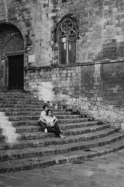 A wide portrait on the stone steps in black and white | Barcelona maternity photoshoot Black-and-white photograph: a wide view of the couple sitting on the steps beneath old stone walls and carved windows during their maternity photoshoot in Barcelona.