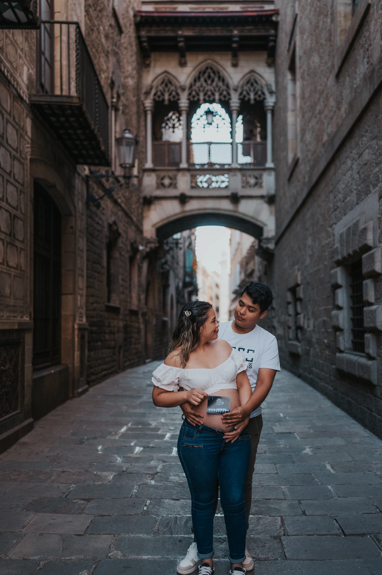 The couple holding an ultrasound photograph together beneath the ornate bridge between the buildings during their maternity photoshoot in Barcelona.