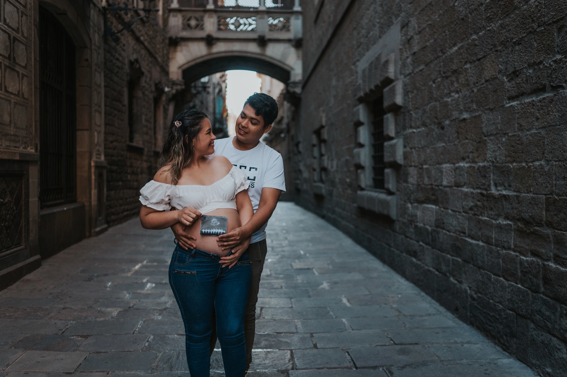 The couple holding an ultrasound photograph together beneath the ornate bridge between the buildings during their maternity photoshoot in Barcelona.