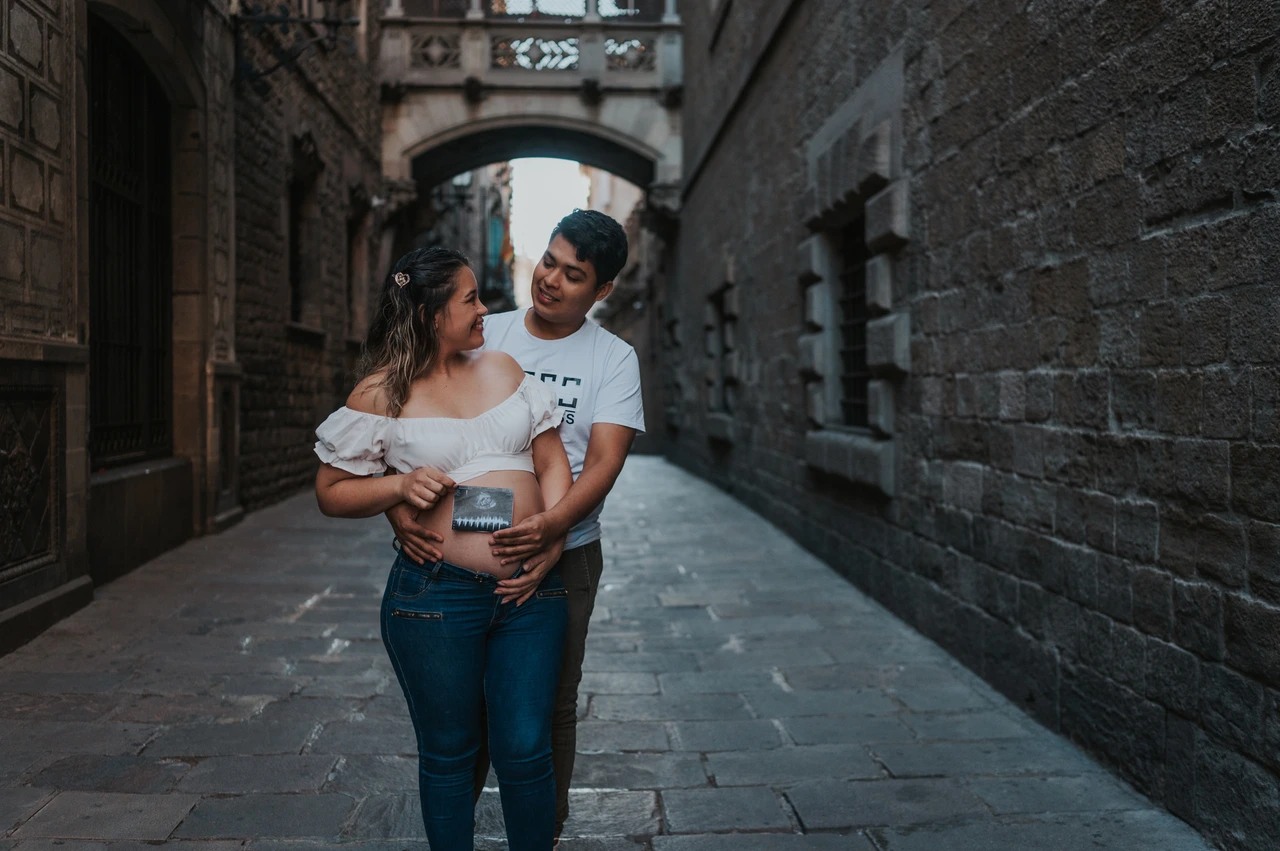Their ultrasound photograph beneath the bridge | Barcelona maternity photoshoot The couple holding an ultrasound photograph together beneath the ornate bridge between the buildings during their maternity photoshoot in Barcelona.