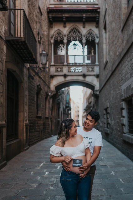 Their ultrasound photograph beneath the bridge | Barcelona maternity photoshoot The couple holding an ultrasound photograph together beneath the ornate bridge between the buildings during their maternity photoshoot in Barcelona.