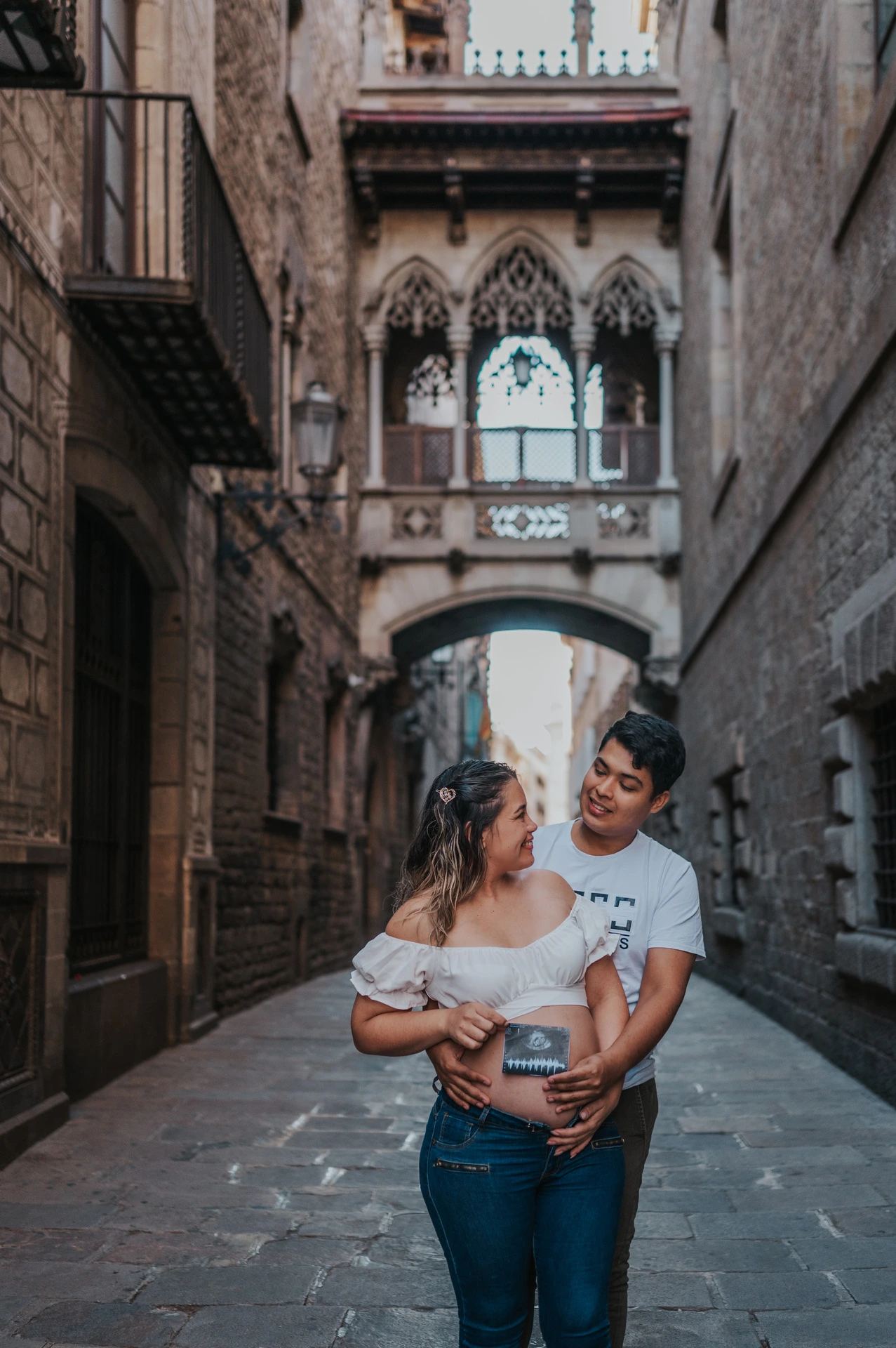 The couple holding an ultrasound photograph together beneath the ornate bridge between the buildings during their maternity photoshoot in Barcelona.