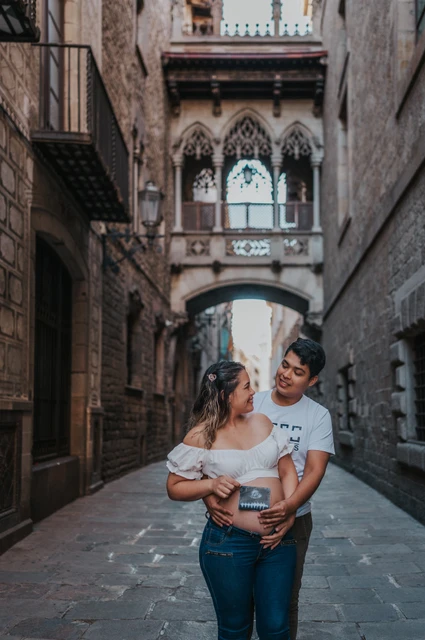 Their ultrasound photograph beneath the bridge | Barcelona maternity photoshoot The couple holding an ultrasound photograph together beneath the ornate bridge between the buildings during their maternity photoshoot in Barcelona.