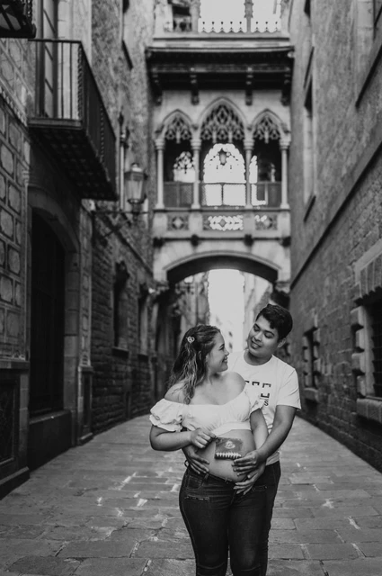 Their ultrasound photograph beneath the bridge in black and white | Barcelona maternity photoshoot Black-and-white photograph: the couple holding an ultrasound photograph together beneath the ornate bridge between the buildings during their maternity photoshoot in Barcelona.