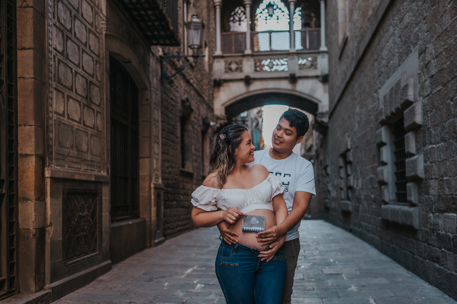 The couple holding an ultrasound photograph together beneath the ornate bridge between the buildings during their maternity photoshoot in Barcelona.
