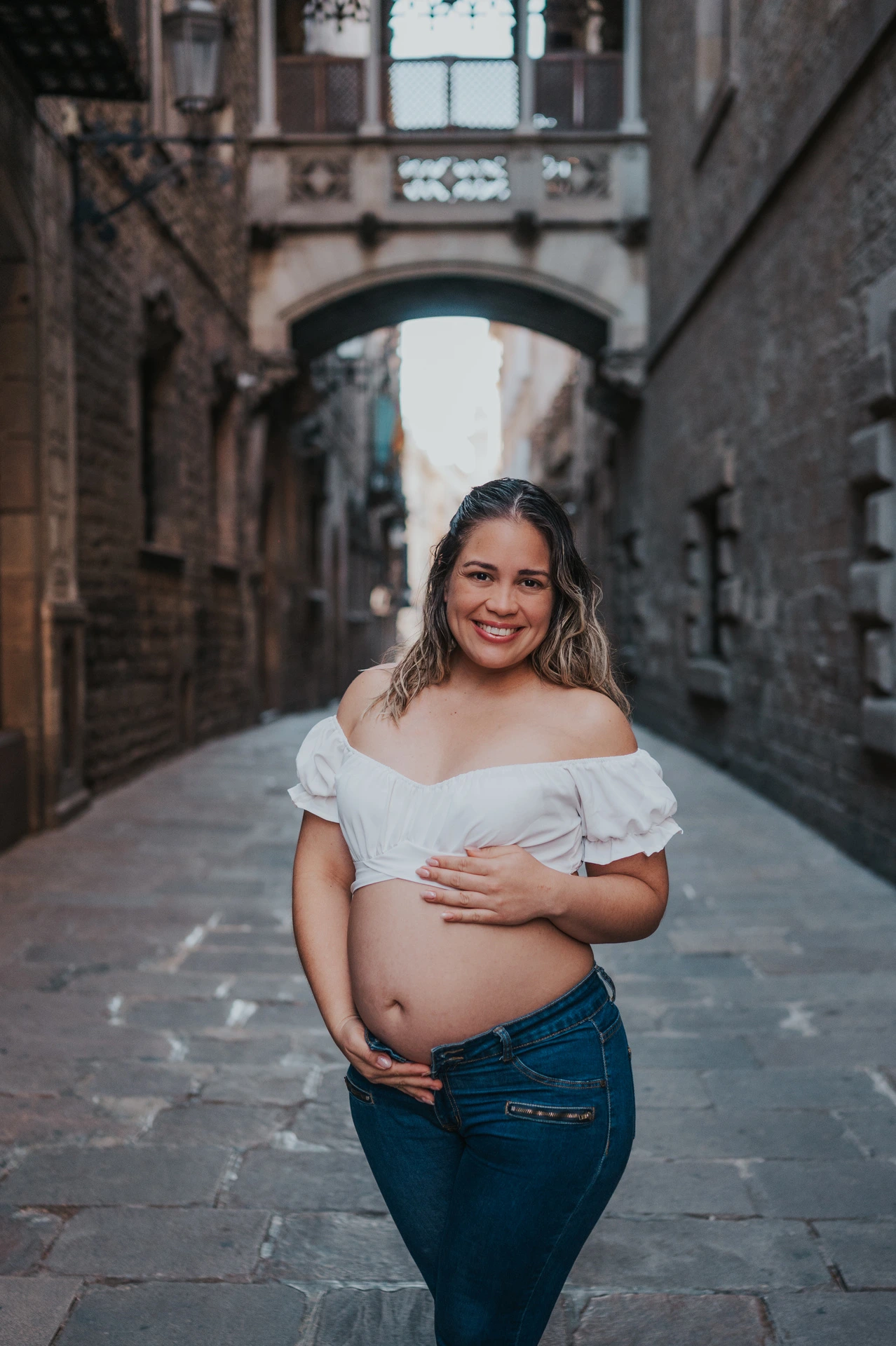 The mother-to-be resting her hands on her bump in the narrow stone street beneath the bridge during their maternity photoshoot in Barcelona.