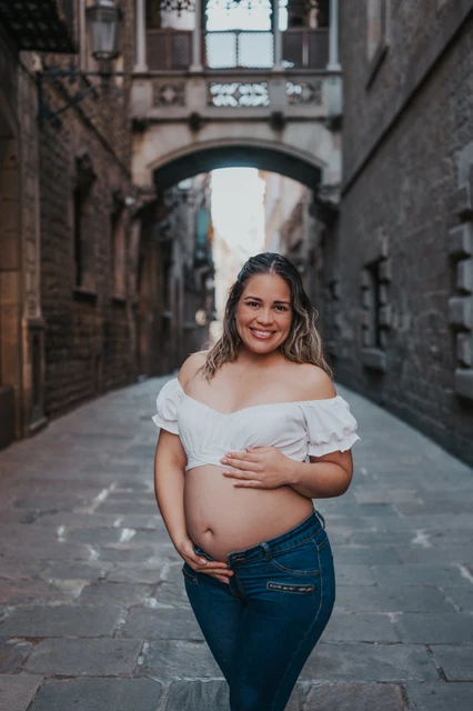A maternity portrait in the narrow stone street | Barcelona maternity photoshoot The mother-to-be resting her hands on her bump in the narrow stone street beneath the bridge during their maternity photoshoot in Barcelona.