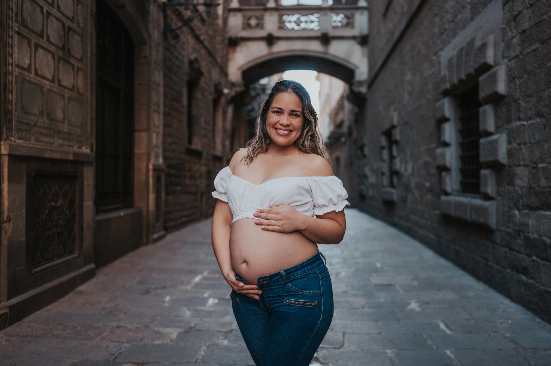 The mother-to-be resting her hands on her bump in the narrow stone street beneath the bridge during their maternity photoshoot in Barcelona.
