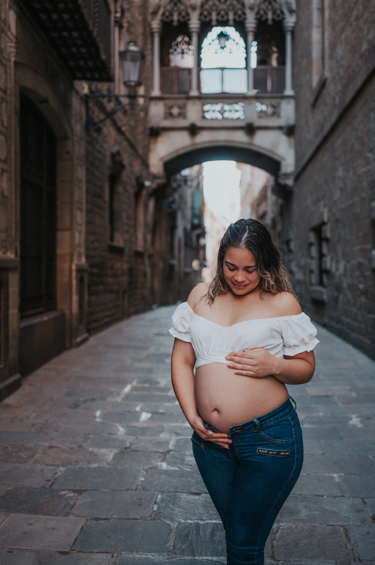 The mother-to-be resting her hands on her bump in the narrow stone street beneath the bridge during their maternity photoshoot in Barcelona.