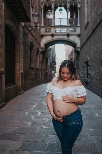 A maternity portrait in the narrow stone street | Barcelona maternity photoshoot The mother-to-be resting her hands on her bump in the narrow stone street beneath the bridge during their maternity photoshoot in Barcelona.