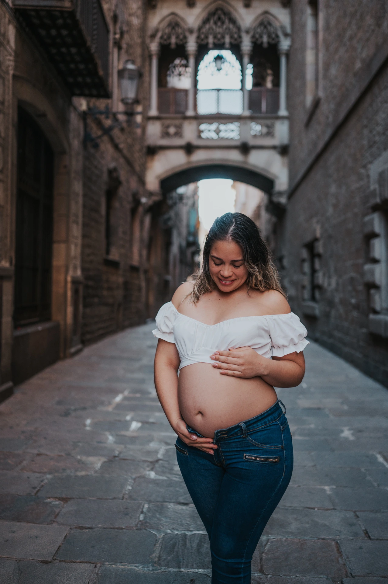 The mother-to-be resting her hands on her bump in the narrow stone street beneath the bridge during their maternity photoshoot in Barcelona.