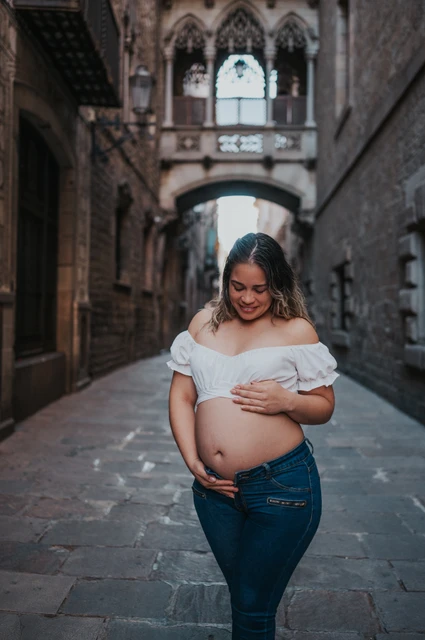 A maternity portrait in the narrow stone street | Barcelona maternity photoshoot The mother-to-be resting her hands on her bump in the narrow stone street beneath the bridge during their maternity photoshoot in Barcelona.