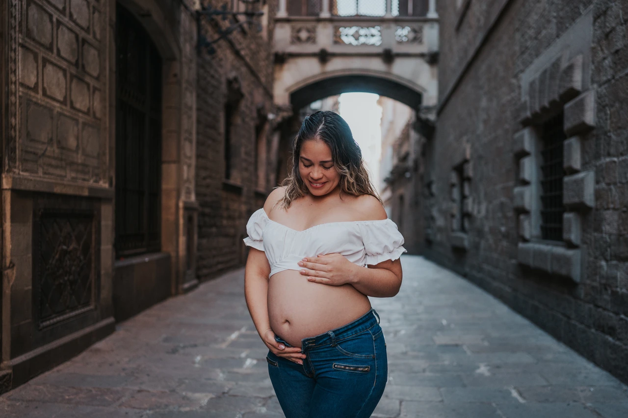A maternity portrait in the narrow stone street | Barcelona maternity photoshoot The mother-to-be resting her hands on her bump in the narrow stone street beneath the bridge during their maternity photoshoot in Barcelona.