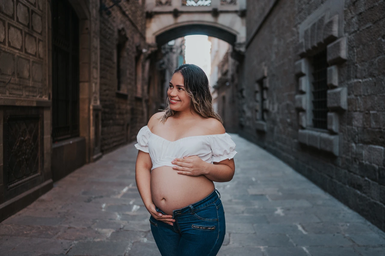 A maternity portrait in the narrow stone street | Barcelona maternity photoshoot The mother-to-be resting her hands on her bump in the narrow stone street beneath the bridge during their maternity photoshoot in Barcelona.