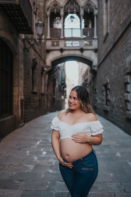 A maternity portrait in the narrow stone street | Barcelona maternity photoshoot The mother-to-be resting her hands on her bump in the narrow stone street beneath the bridge during their maternity photoshoot in Barcelona.