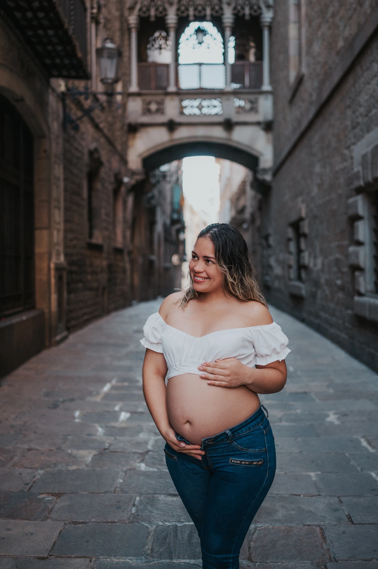 The mother-to-be resting her hands on her bump in the narrow stone street beneath the bridge during their maternity photoshoot in Barcelona.