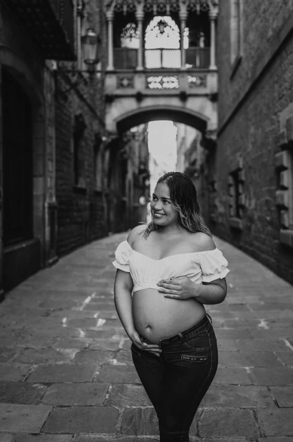 A maternity portrait in the narrow stone street in black and white | Barcelona maternity photoshoot Black-and-white photograph: the mother-to-be resting her hands on her bump in the narrow stone street beneath the bridge during their maternity photoshoot in Barcelona.