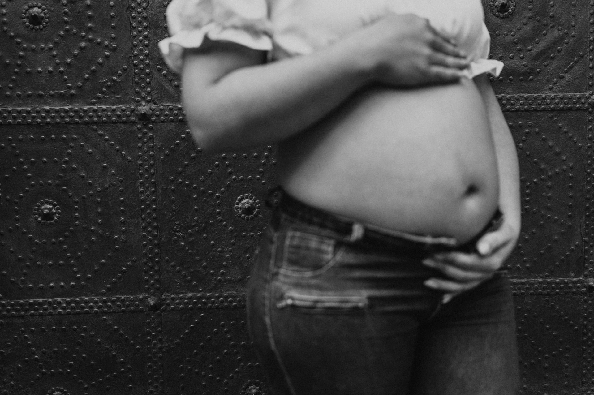 Black-and-white photograph: a close view of the mother-to-be’s hands resting on her bump against a dark door during their maternity photoshoot in Barcelona.