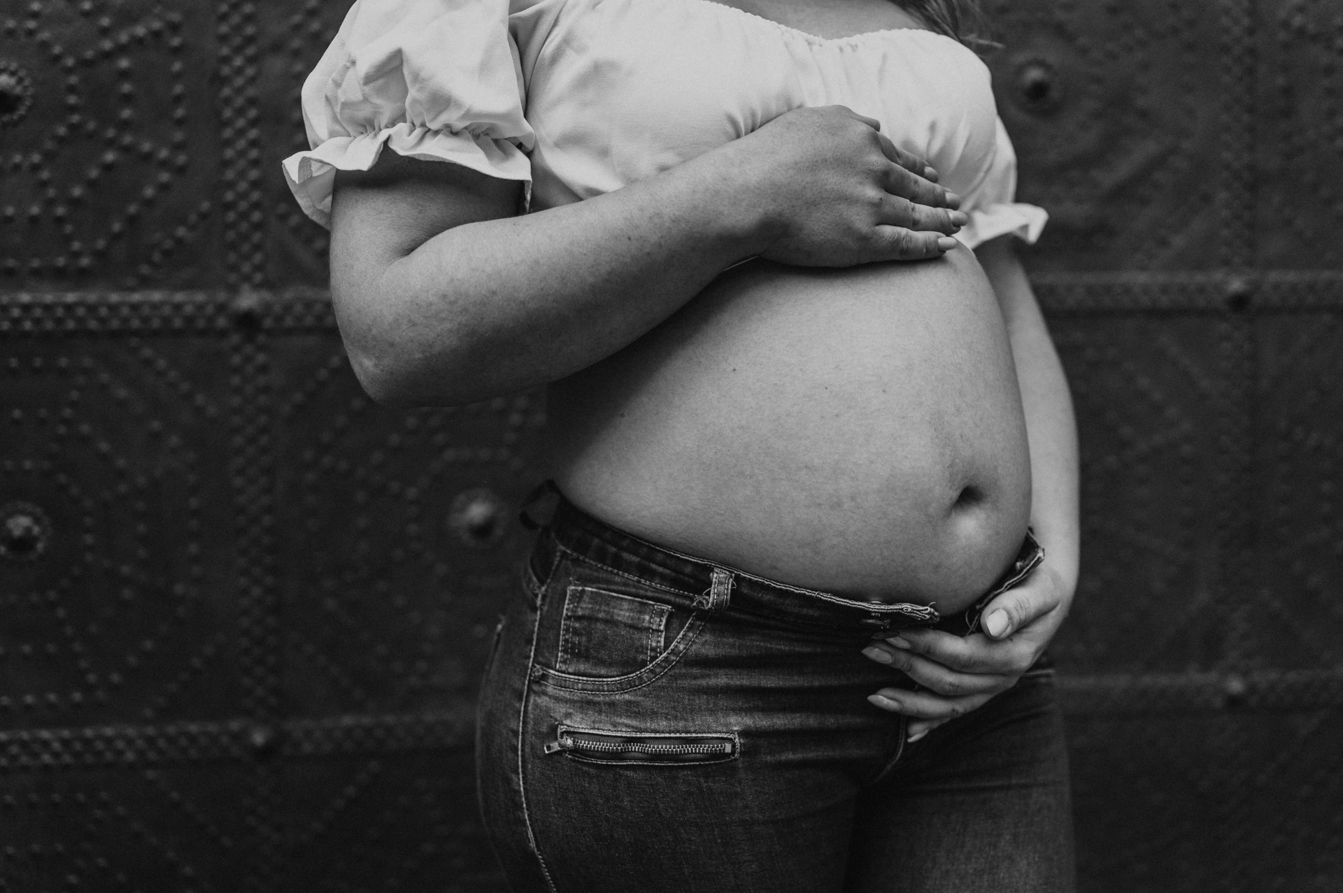 Black-and-white photograph: a close view of the mother-to-be’s hands resting on her bump against a dark door during their maternity photoshoot in Barcelona.