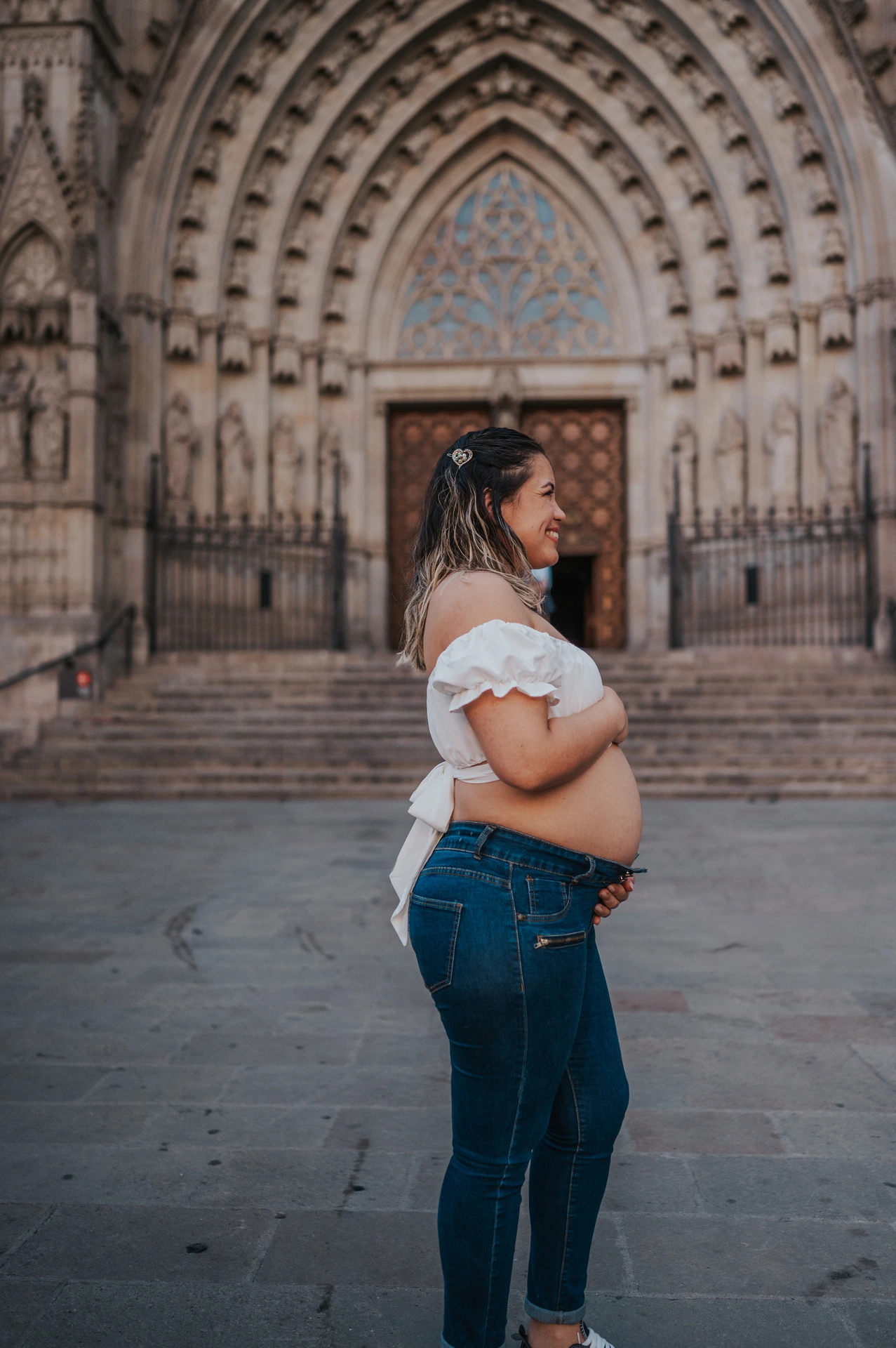 The mother-to-be standing sideways with her hands on her bump in front of the cathedral entrance during their maternity photoshoot in Barcelona.