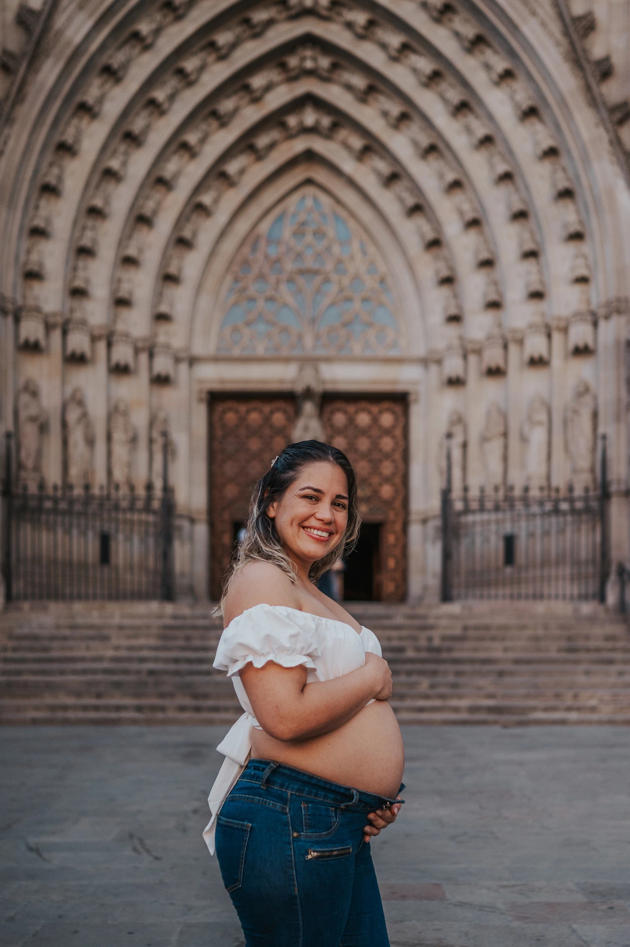 The mother-to-be standing sideways with her hands on her bump in front of the cathedral entrance during their maternity photoshoot in Barcelona.