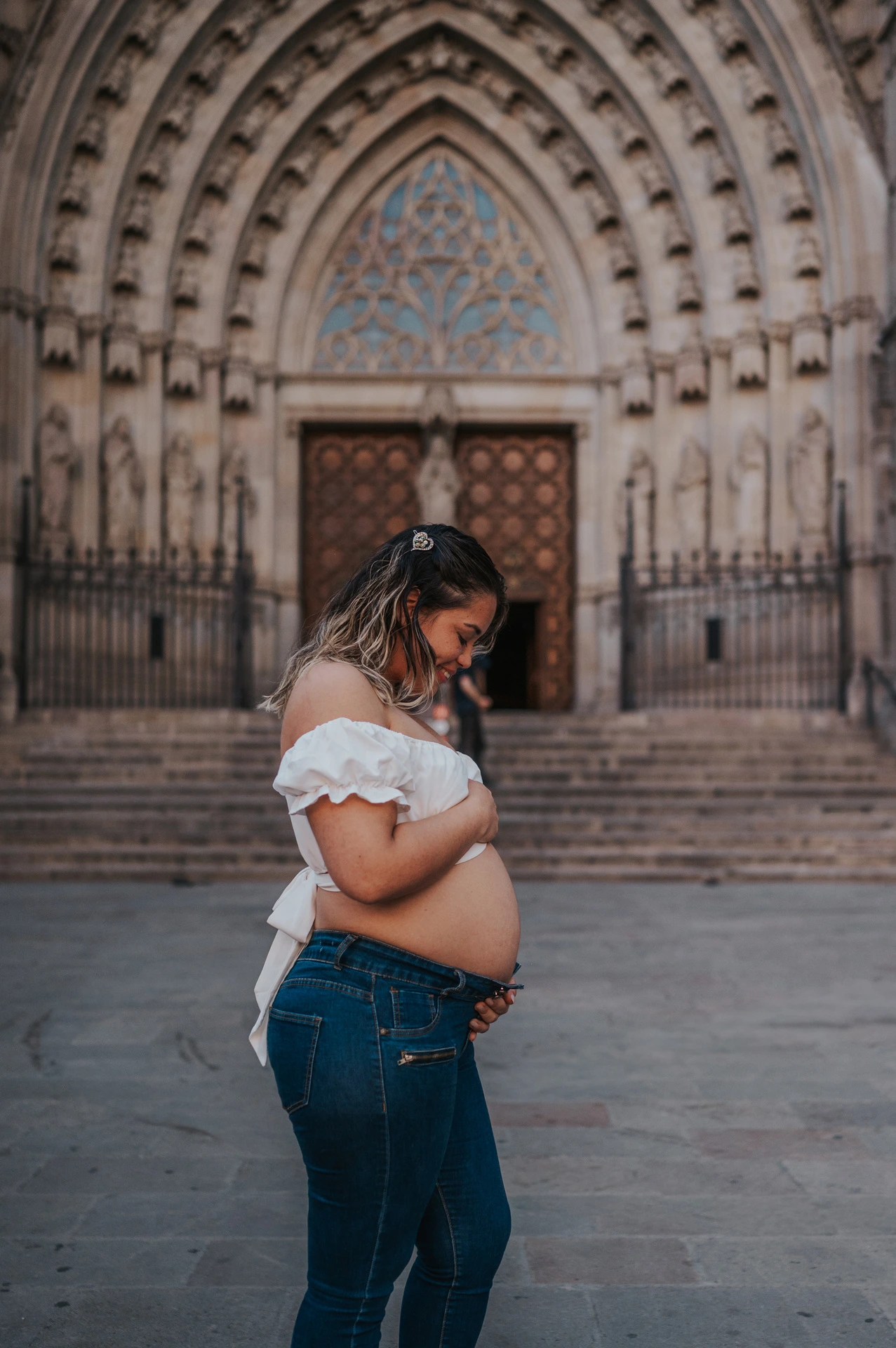 The mother-to-be standing sideways with her hands on her bump in front of the cathedral entrance during their maternity photoshoot in Barcelona.