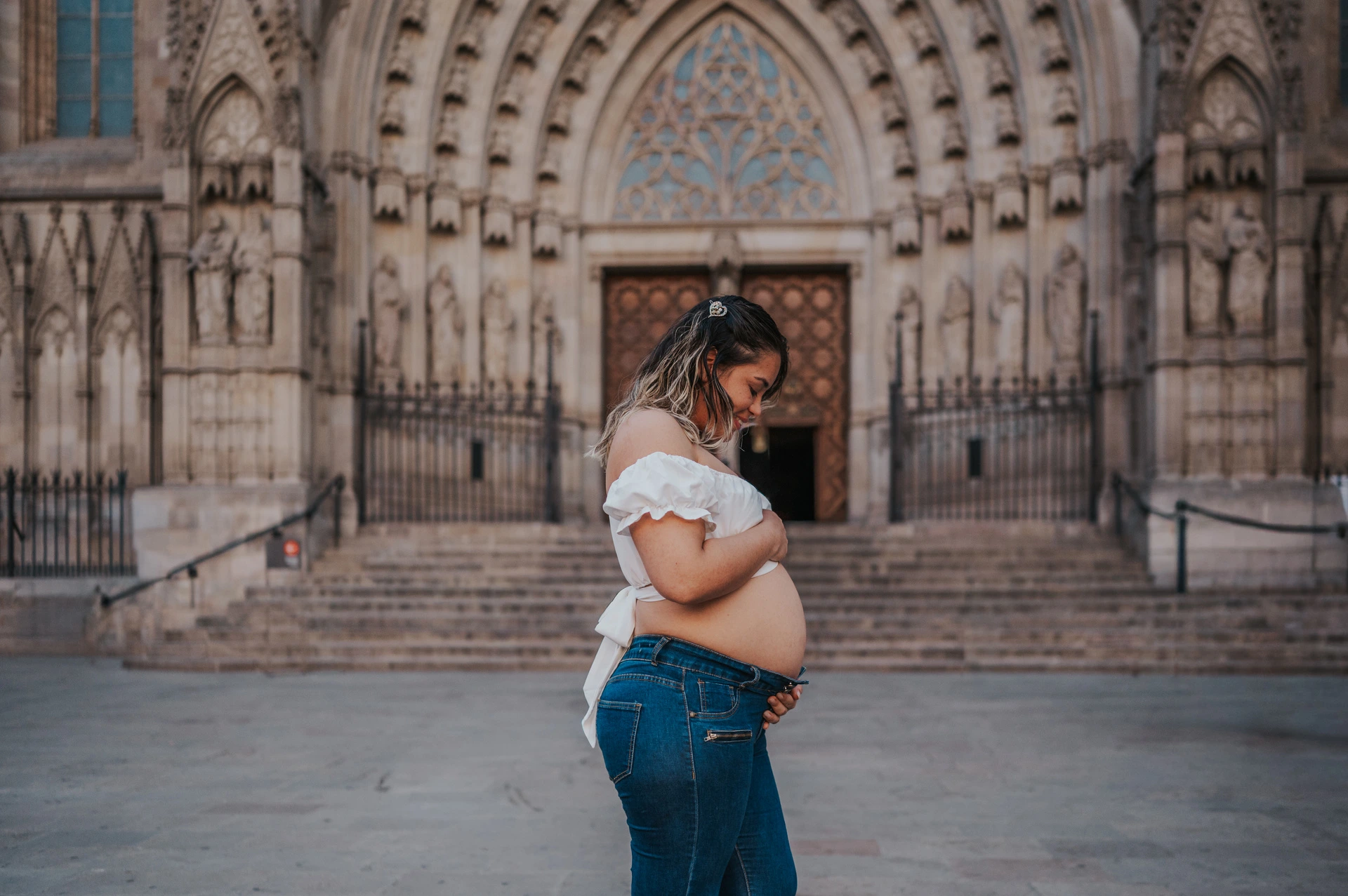 The mother-to-be standing sideways with her hands on her bump in front of the cathedral entrance during their maternity photoshoot in Barcelona.