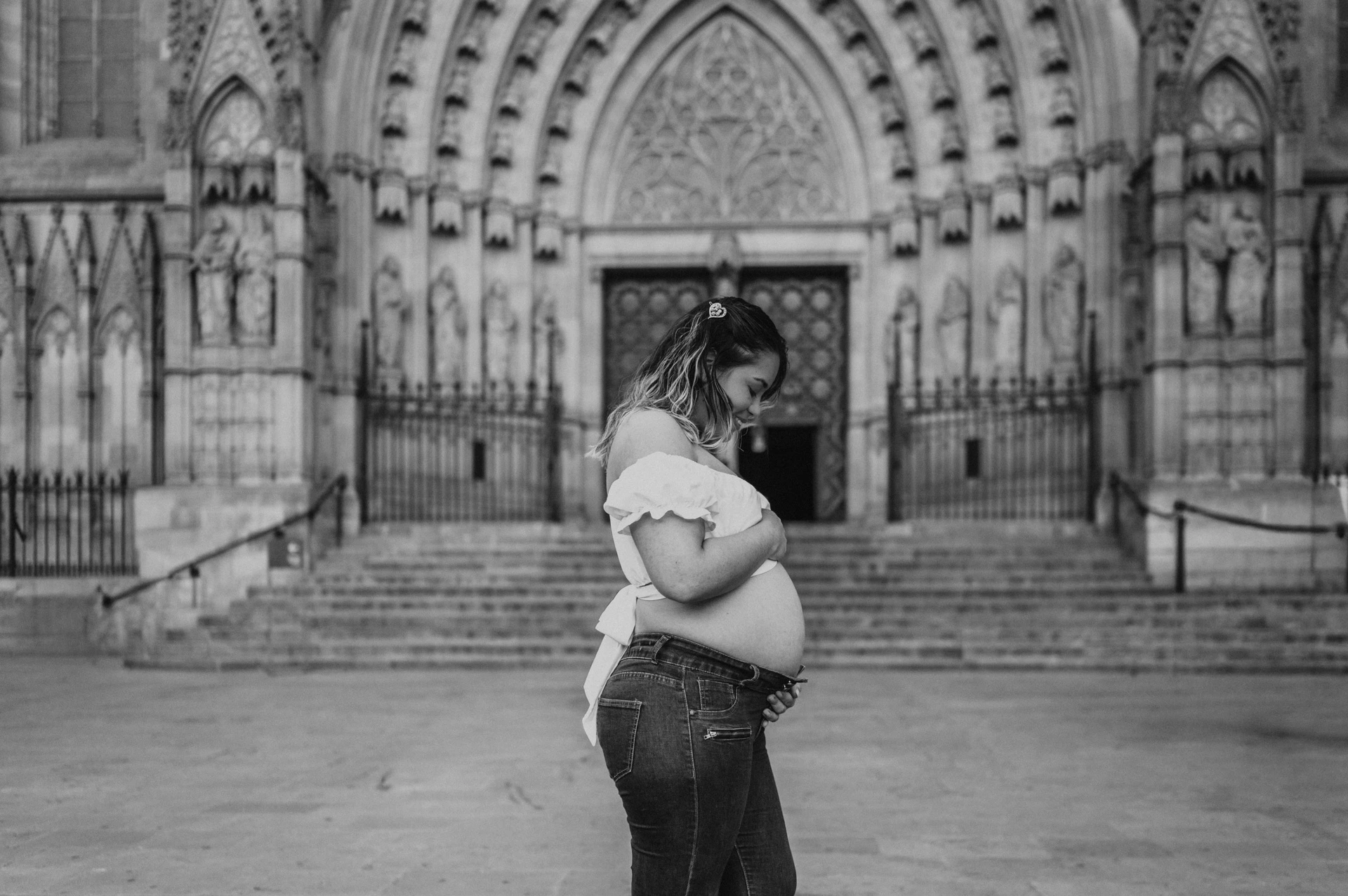 Black-and-white photograph: the mother-to-be standing sideways with her hands on her bump in front of the cathedral entrance during their maternity photoshoot in Barcelona.