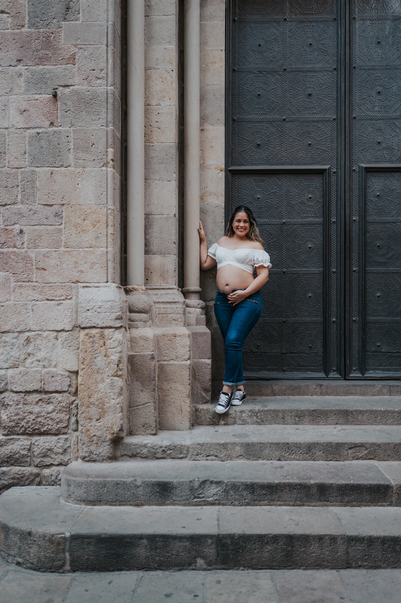 The mother-to-be holding her bump beside the large doorway and stone steps during their maternity photoshoot in Barcelona.