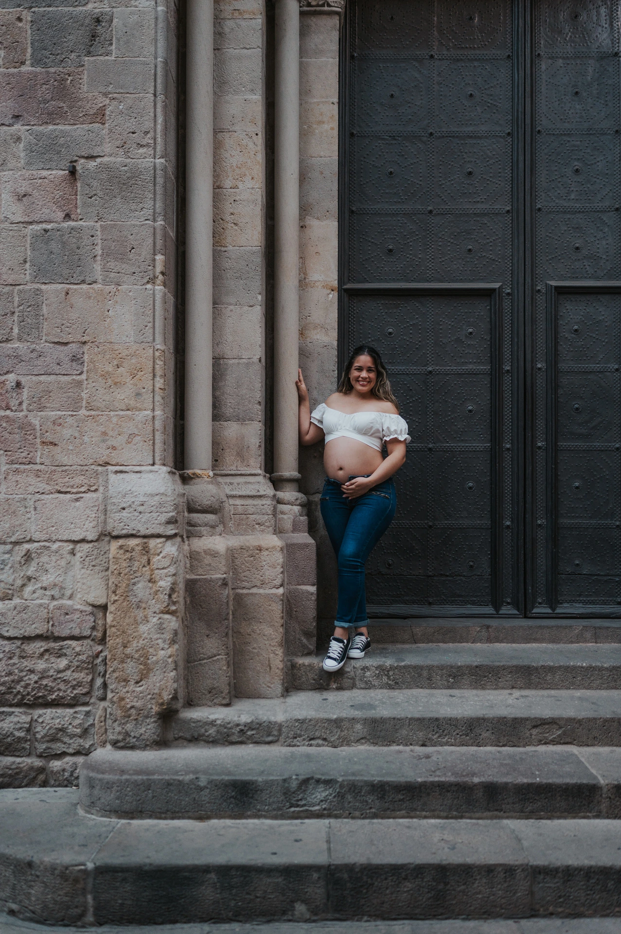 The mother-to-be holding her bump beside the large doorway and stone steps during their maternity photoshoot in Barcelona.