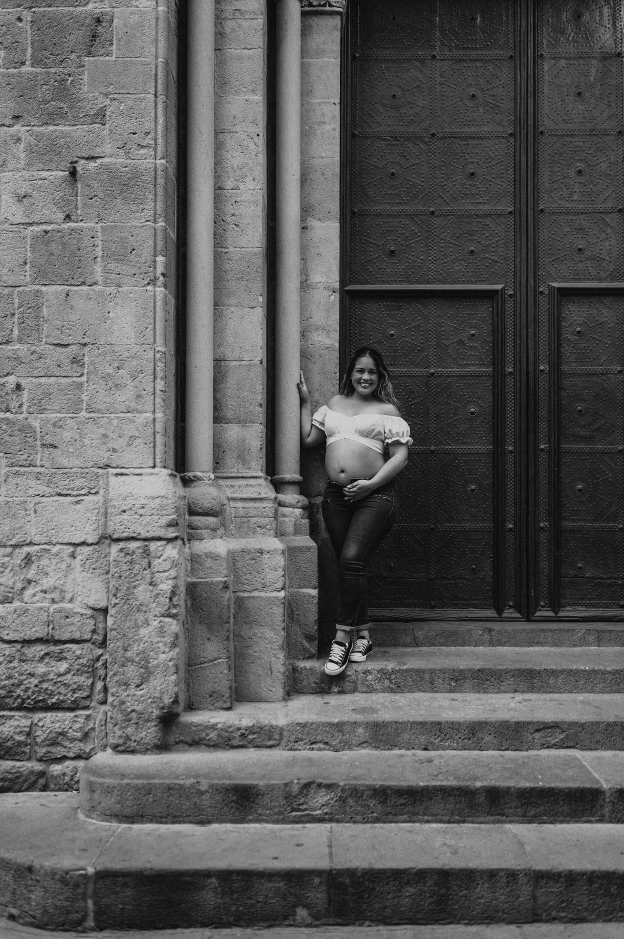 Black-and-white photograph: the mother-to-be holding her bump beside the large doorway and stone steps during their maternity photoshoot in Barcelona.