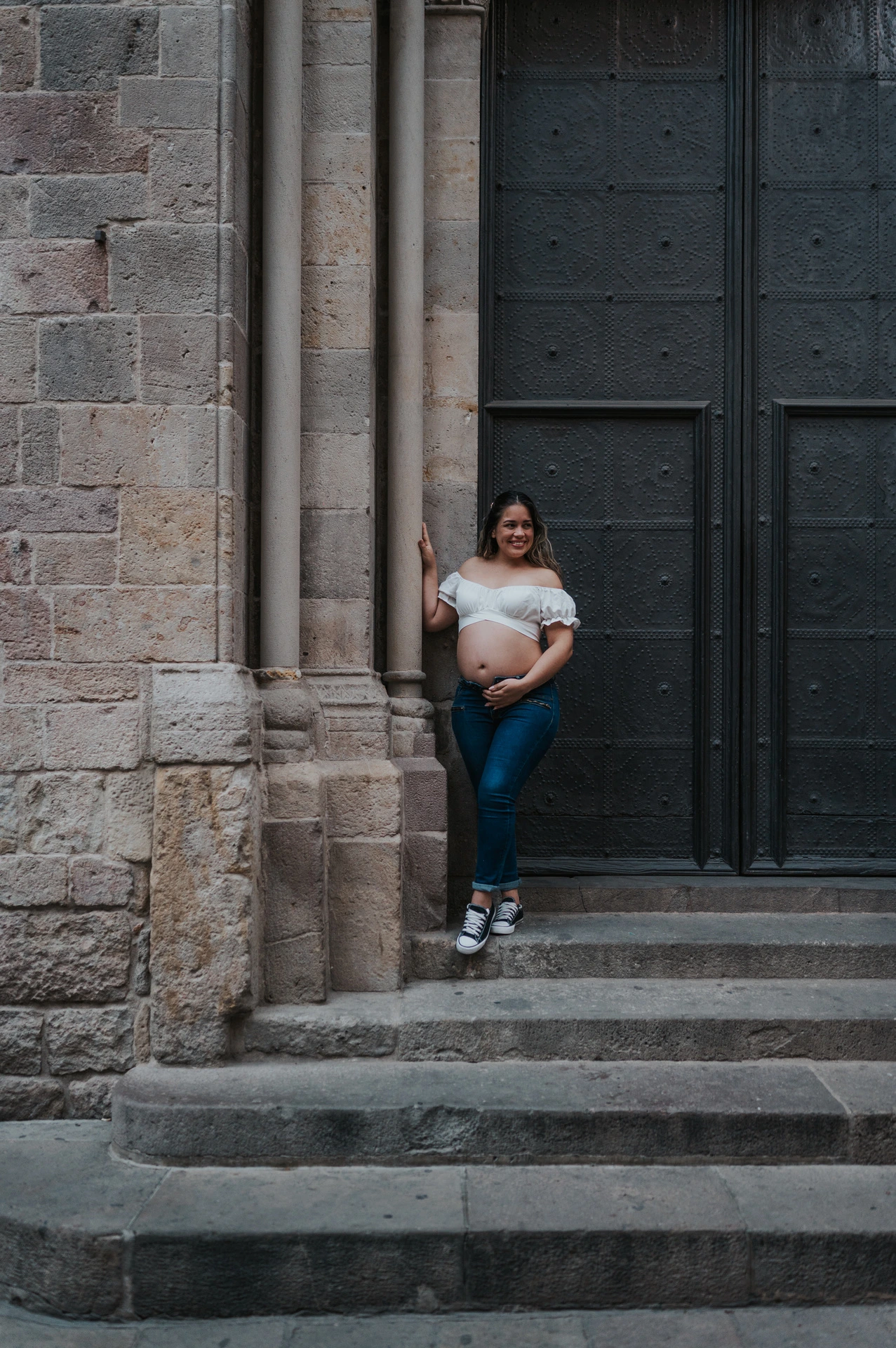 The mother-to-be holding her bump beside the large doorway and stone steps during their maternity photoshoot in Barcelona.
