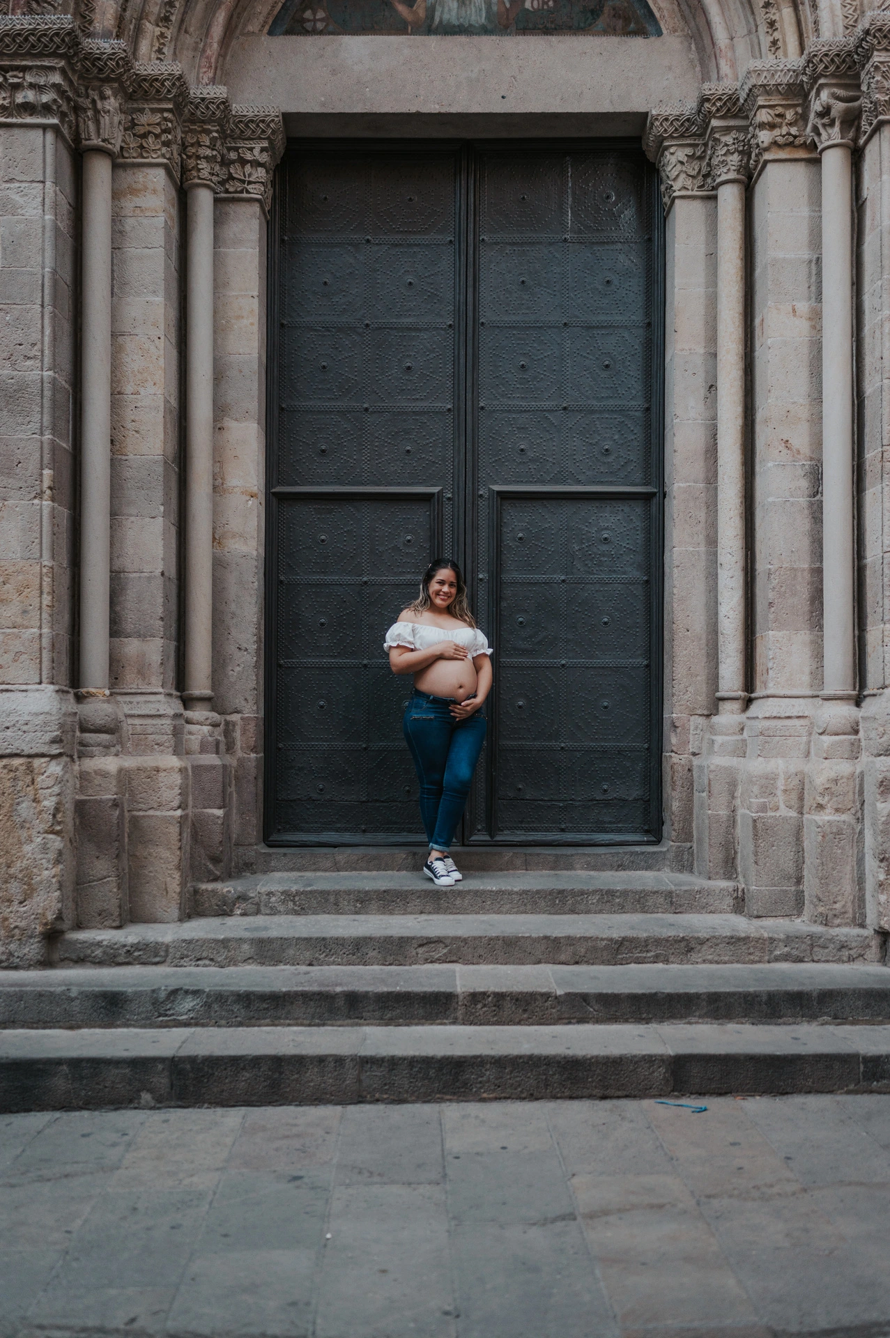 The mother-to-be holding her bump beside the large doorway and stone steps during their maternity photoshoot in Barcelona.