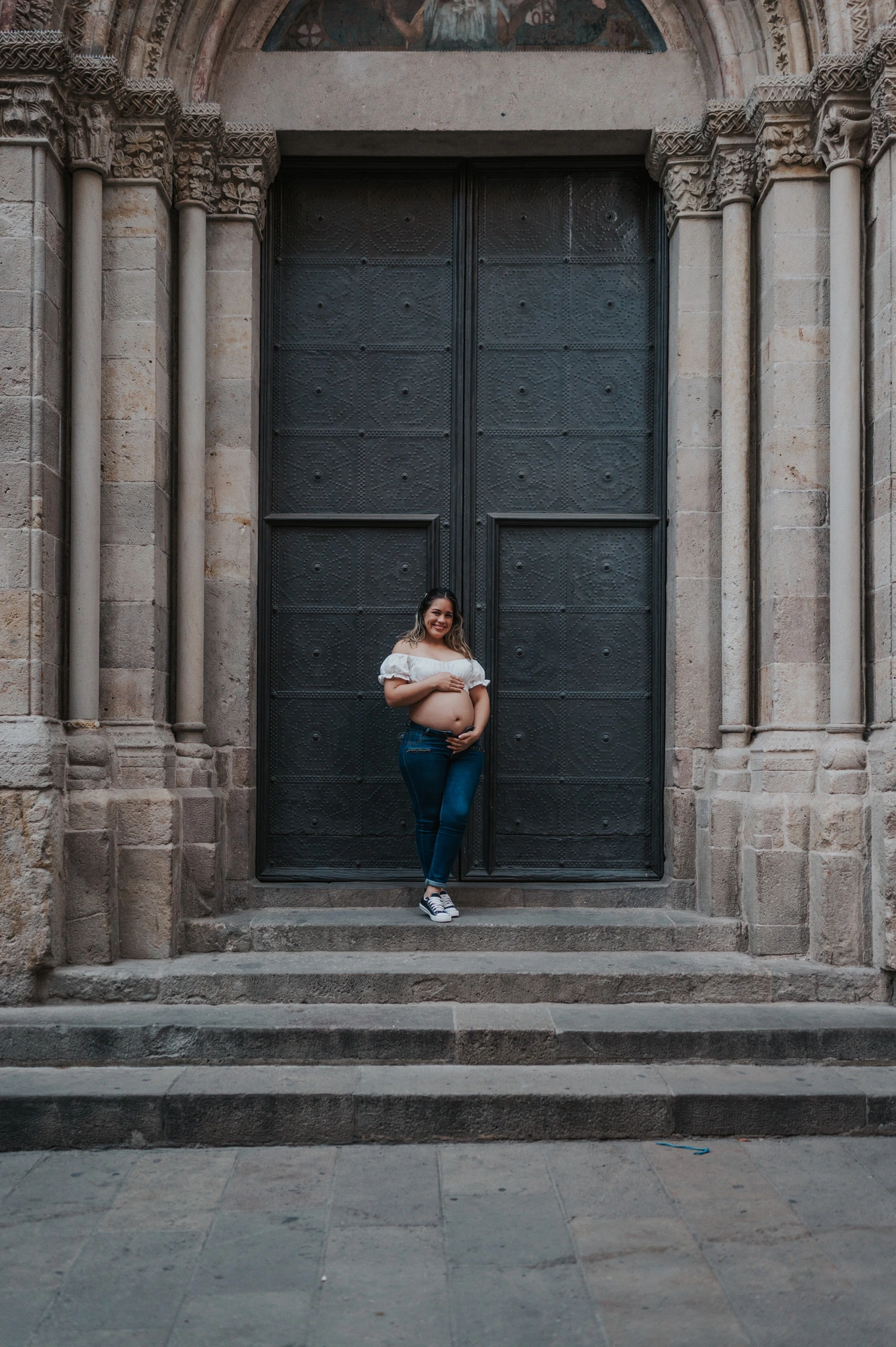 The mother-to-be holding her bump beside the large doorway and stone steps during their maternity photoshoot in Barcelona.