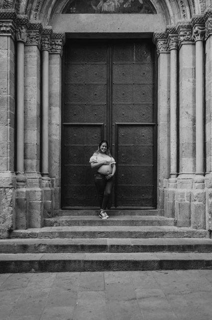A maternity portrait beside the stone doorway in black and white | Barcelona maternity photoshoot Black-and-white photograph: the mother-to-be holding her bump beside the large doorway and stone steps during their maternity photoshoot in Barcelona.