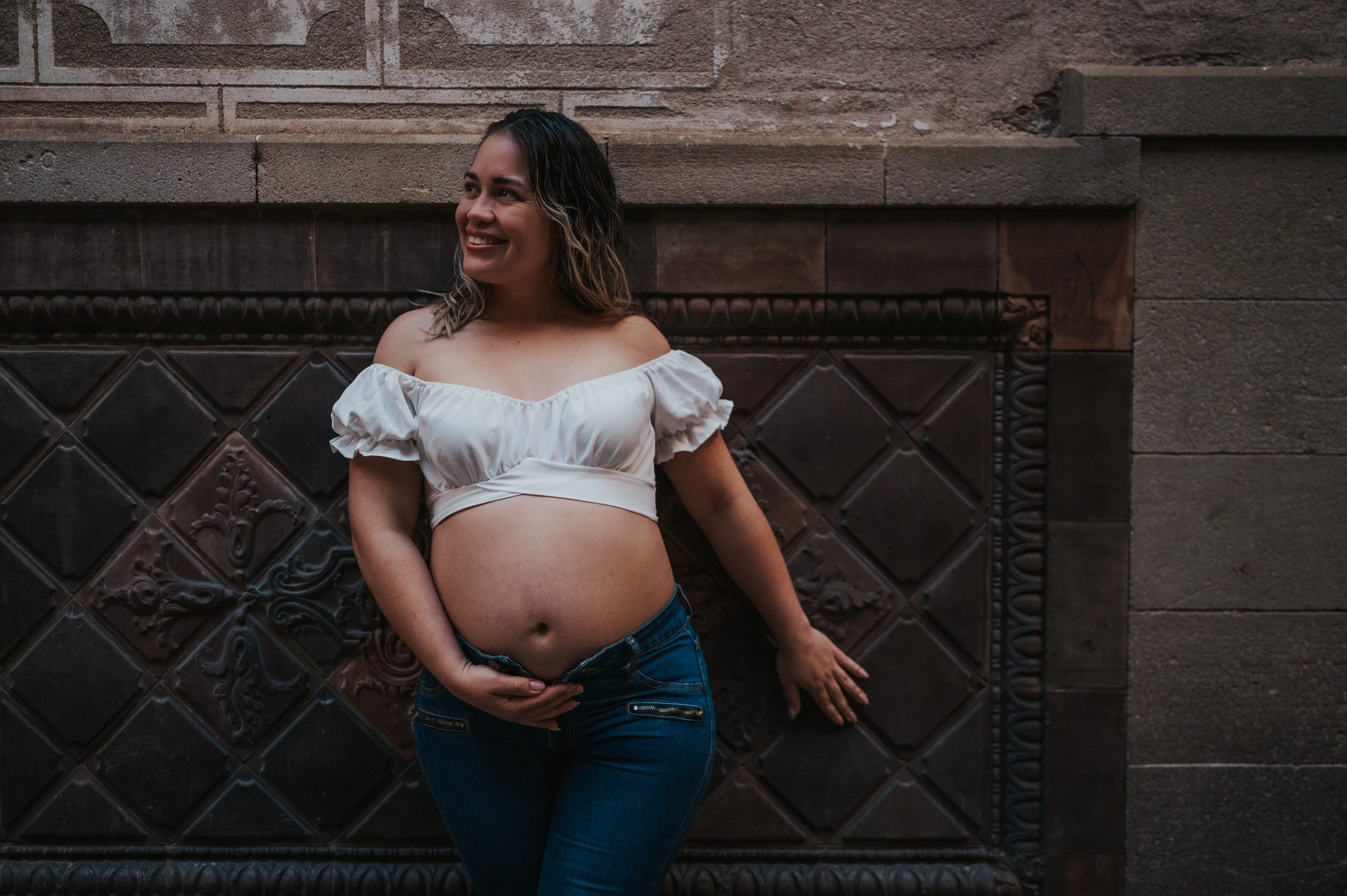 The mother-to-be standing beside a patterned wall with her hand resting on her bump during their maternity photoshoot in Barcelona.