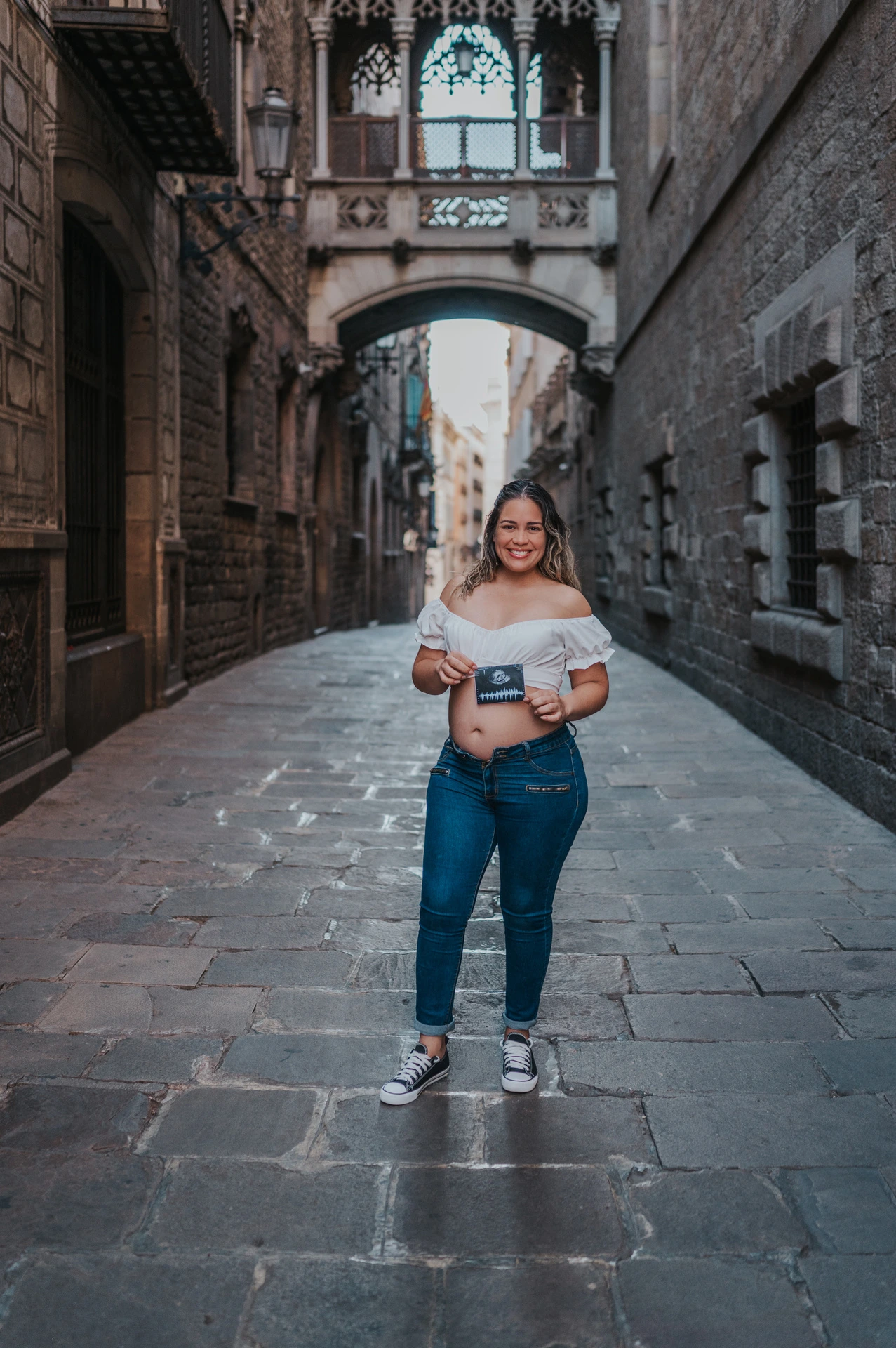 The mother-to-be holding an ultrasound photograph in front of her bump beneath the ornate bridge during their maternity photoshoot in Barcelona.