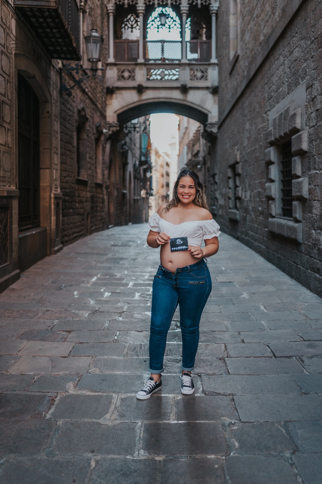 The mother-to-be holding an ultrasound photograph in front of her bump beneath the ornate bridge during their maternity photoshoot in Barcelona.