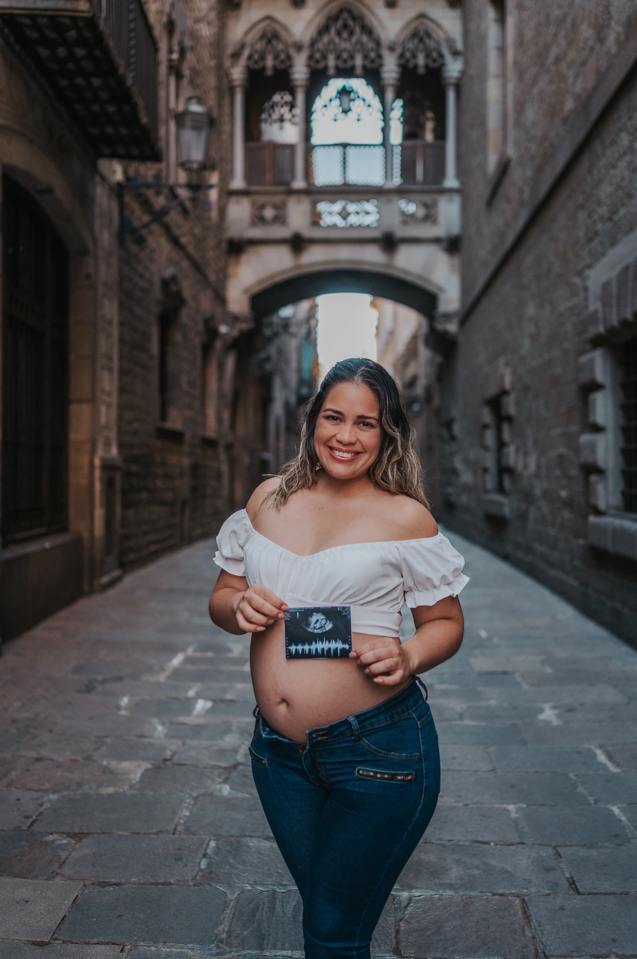 The mother-to-be holding an ultrasound photograph in front of her bump beneath the ornate bridge during their maternity photoshoot in Barcelona.