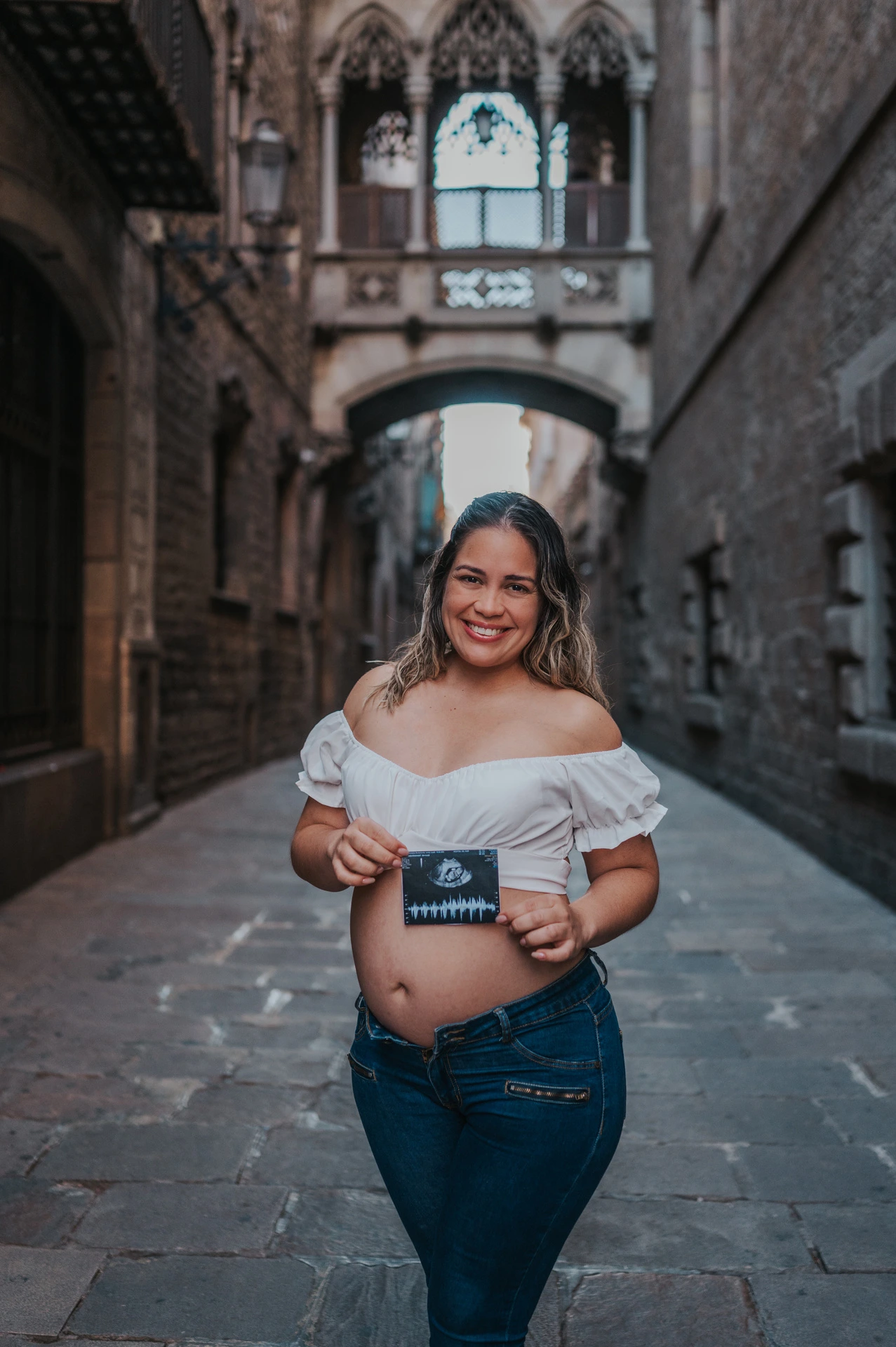 The mother-to-be holding an ultrasound photograph in front of her bump beneath the ornate bridge during their maternity photoshoot in Barcelona.