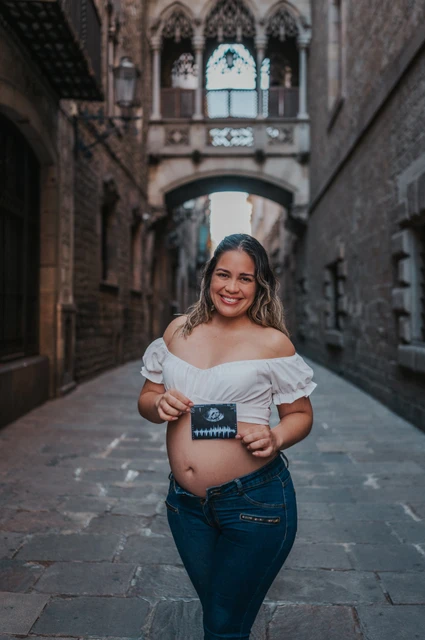The mother-to-be holding an ultrasound photograph in front of her bump beneath the ornate bridge during their maternity photoshoot in Barcelona.