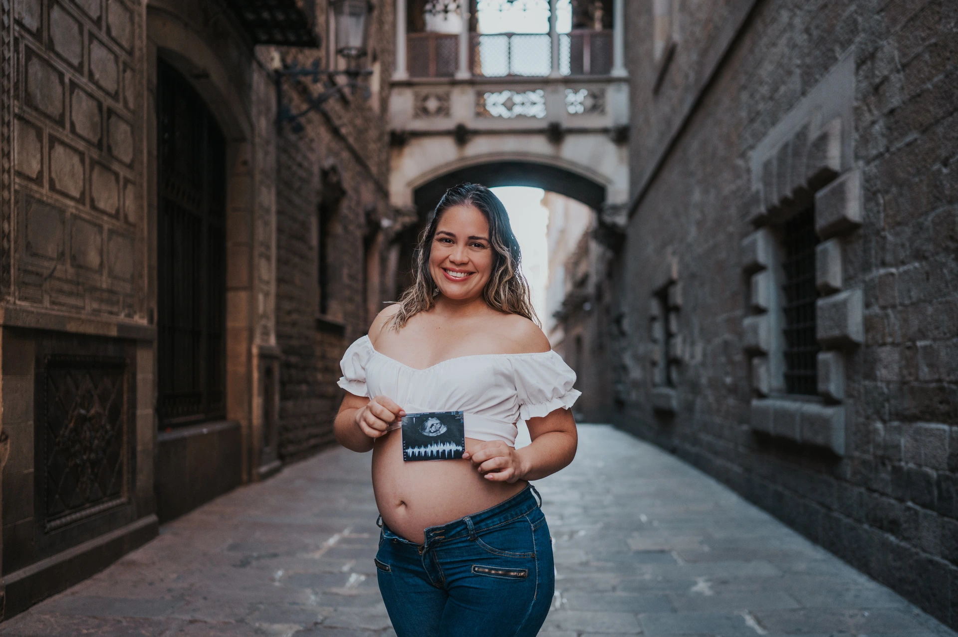 The mother-to-be holding an ultrasound photograph in front of her bump beneath the ornate bridge during their maternity photoshoot in Barcelona.