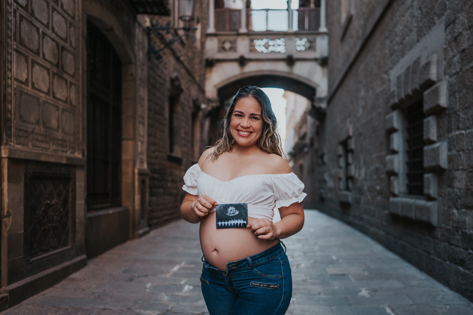 The mother-to-be holding an ultrasound photograph in front of her bump beneath the ornate bridge during their maternity photoshoot in Barcelona.