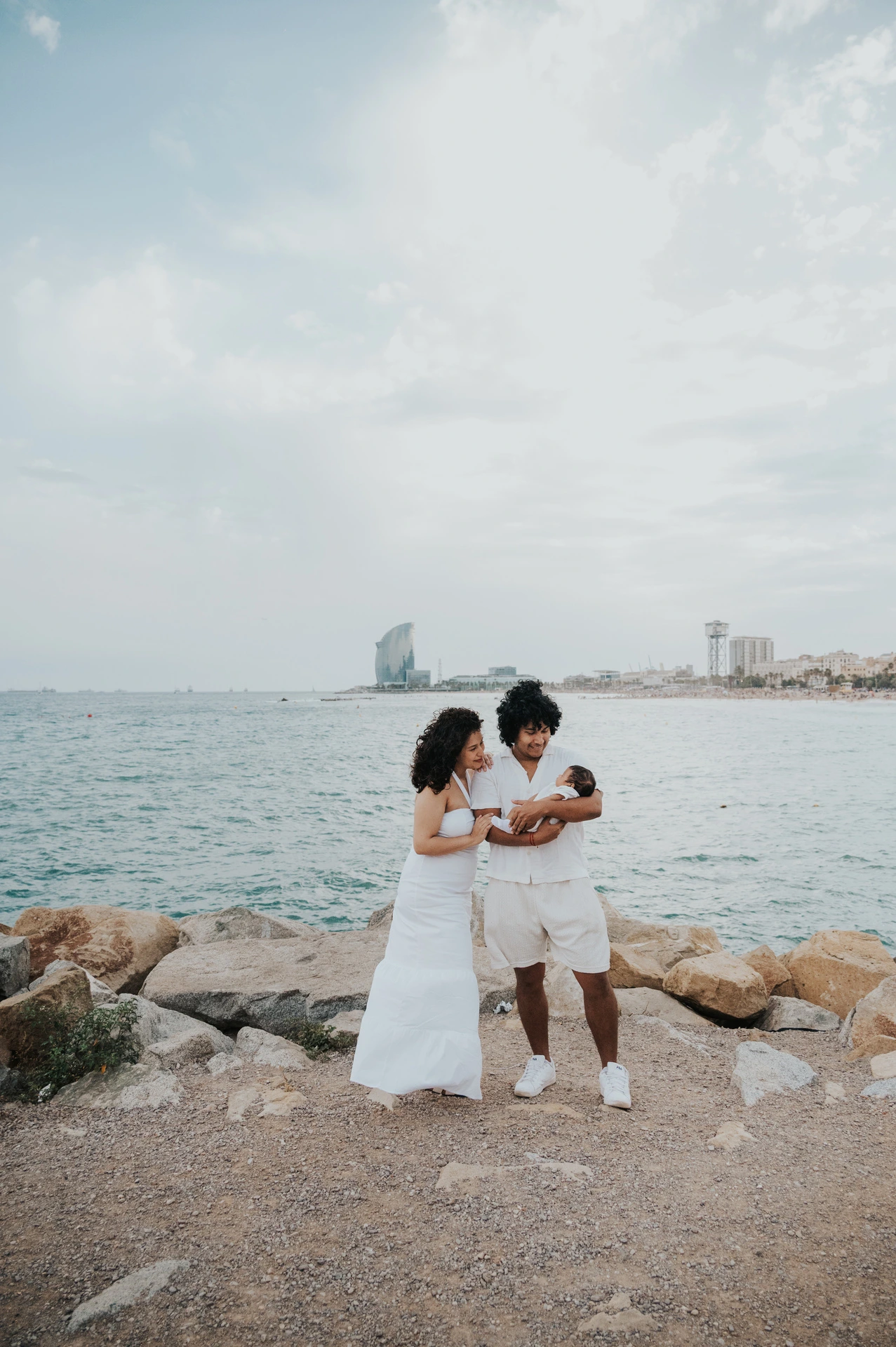 The parents holding their newborn beside the shoreline with the city across the water during their newborn family photoshoot in Barcelona.