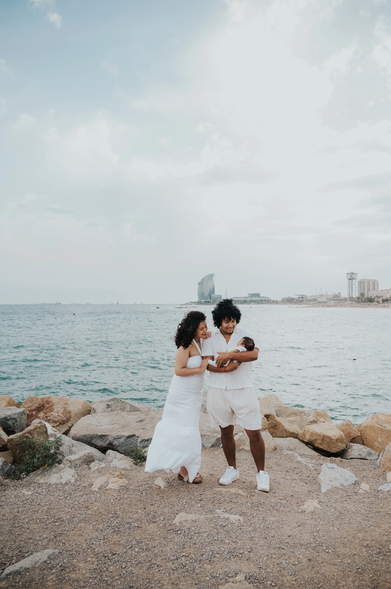 The parents holding their newborn beside the shoreline with the city across the water during their newborn family photoshoot in Barcelona.