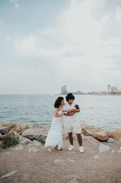 A wide family portrait beside the sea | Barcelona newborn family photoshoot The parents holding their newborn beside the shoreline with the city across the water during their newborn family photoshoot in Barcelona.